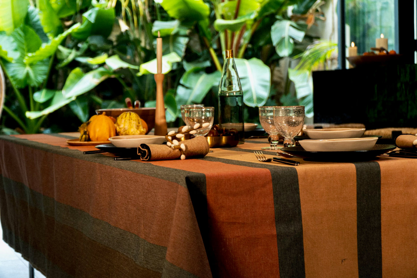 Dining setting with palm trees in the background. A Tablecloth in 100% cotton fabric roll by Les Toiles du Soleil. Automne multi coloured stripes of terracotta, peach, taupe and beige