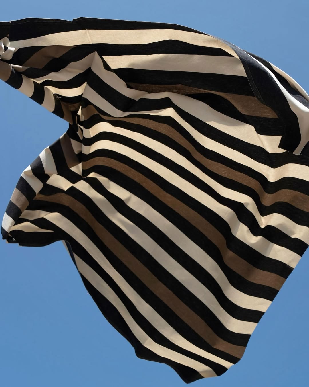 Boulevard Raspail Taupe 100% cotton Les Toiles du Soleil 180cm with black, brown and neutral multi striped fabric lifestyle shot of fabric being thrown in the air with sky blue background