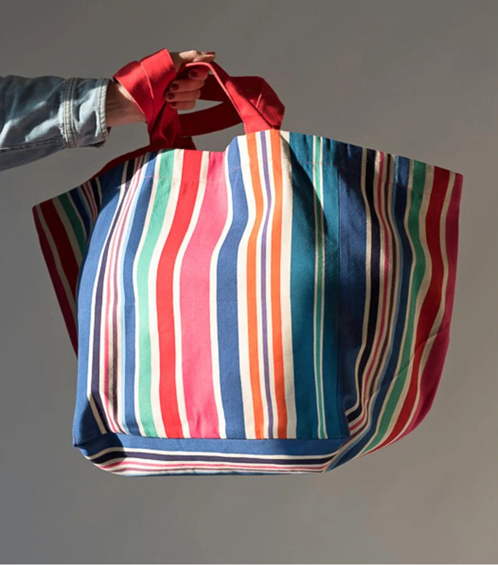 Product photoshoot of a hand holding a large carry bag made from Les Toiles du Soleil Cabanon fabric - featuring bold stripes of navy, red, fuchsia, orange, teal, green, cream and purple, with a fine orange selvedge edge.