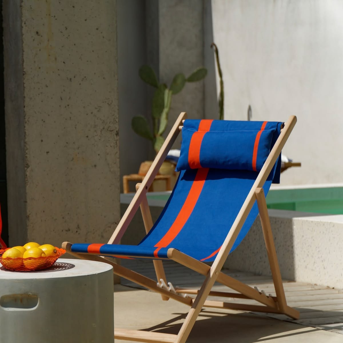 Home pool area with minimalist design of concrete. Featuring a deckchair with Rue du Soleil cotton fabric roll in vivid cobalt blue with fine bright orange accent stripes. Made by Les Toiles do Soleil.