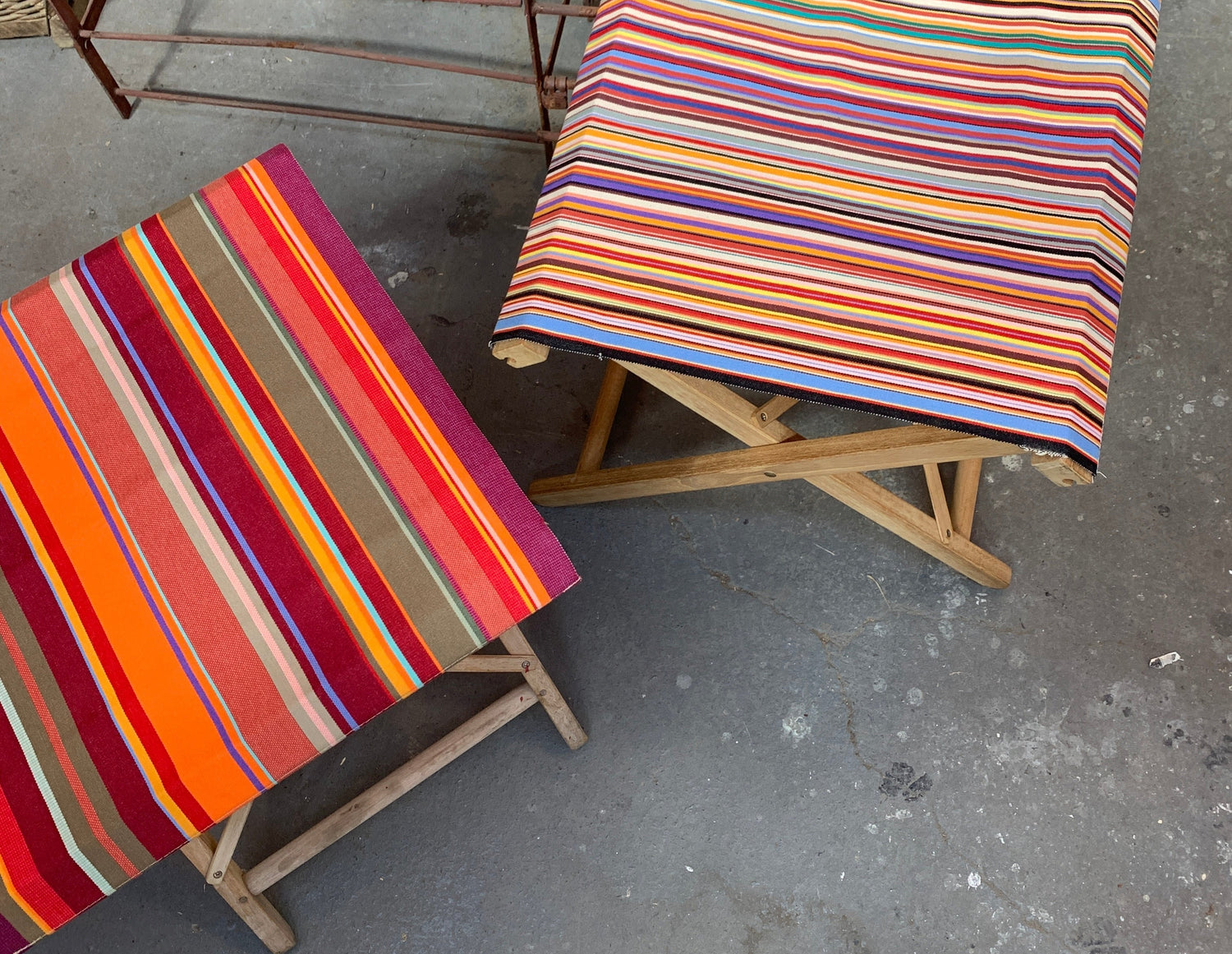 Two wooden stools and fisherman chairs with colourful Sunbrella and Lestoiles du Soleil striped covers on a concrete floor.