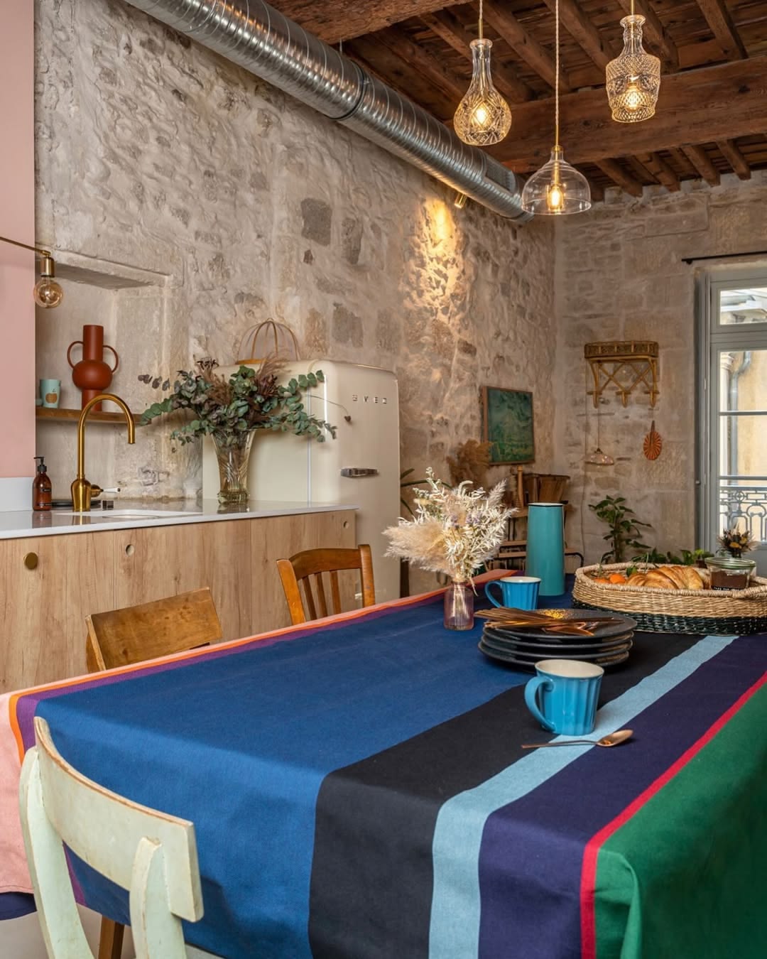 Dining room set-up in a rustic room with fabric Tablecloth of Lucas - a fabric bold striped Les Toiles du Soleil woven fabric of deep purple, blush pink, burnt orange, petrol blue, black and forest green. The tablecloth has plates, cutlery and a basket of fruit and pastries on it