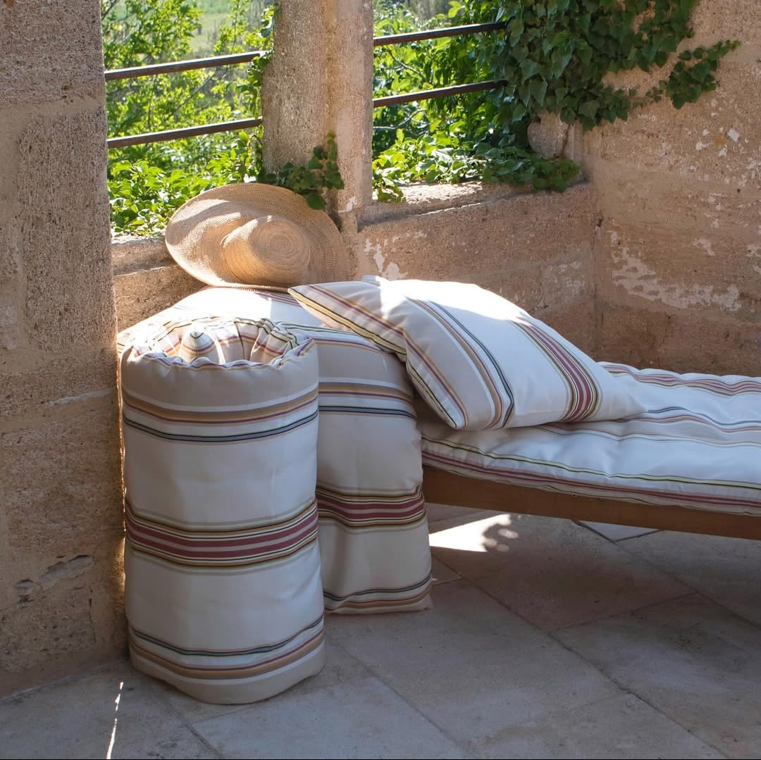 Outdoor seating area with a Les Toiles du Soleil cushioned bench and ottoman against a stone wall with greenery.