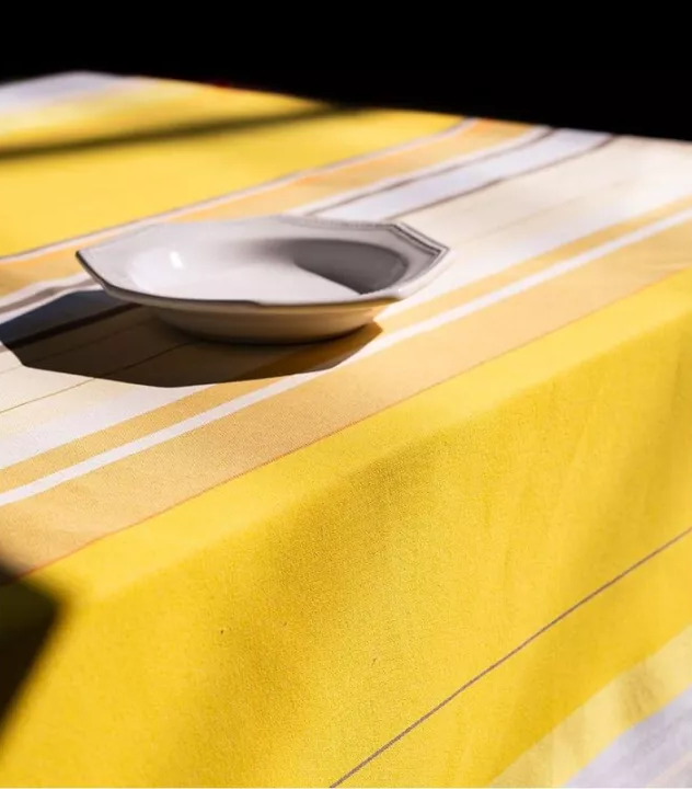 Table cloth of Plein Soleil multi coloured yellow, grey, and sun toned striped Les Toiles du Soleil 100% cotton fabric. With white ceramic bowl placed on top