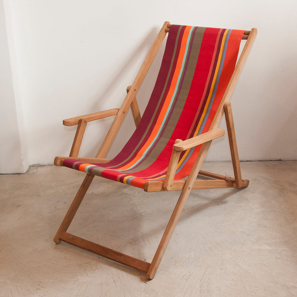 Ici et la teak deckchair with arms made from Collioure Rouge - a fabric featuring a striped pattern with various bands in reds, orange, taupe, burgundy, turquoise, and other vibrant colours. A detailed 100% cotton weave by By Les Toiles du Soleil