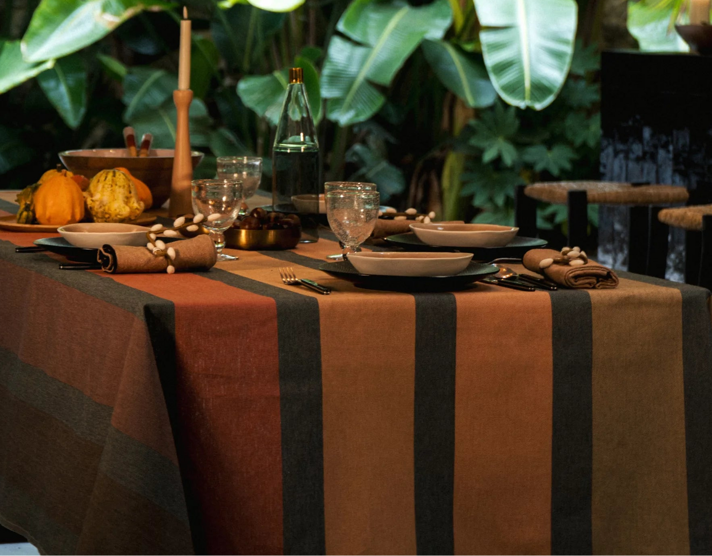 Dining setting with palm trees in the background. A Tablecloth in 100% cotton fabric roll by Les Toiles du Soleil. Automne multi coloured stripes of terracotta, peach, taupe and beige