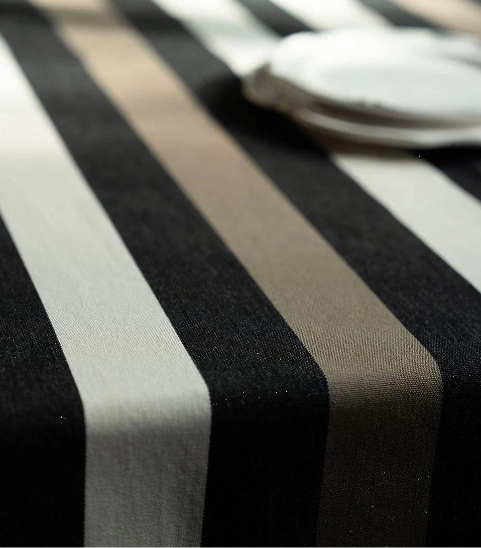 Table cloth from Boulevard Raspail Taupe 100% cotton Les Toiles du Soleil 180cm with black, brown and neutral multi striped  fabric with two ceramic plates blurred in background