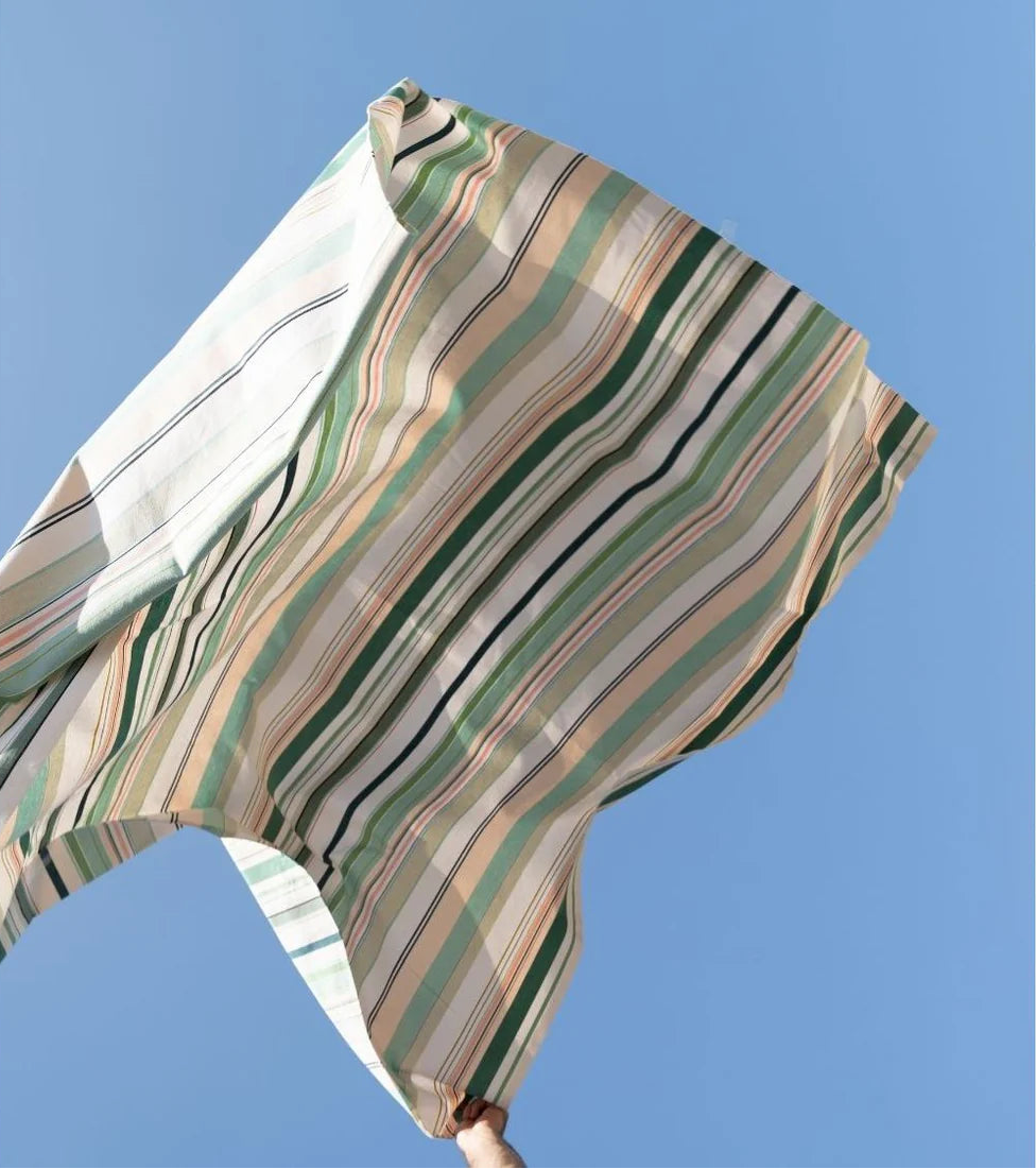 Les Toiles du soleil Bompas fabric thrown in the air with sky blue background. The  multi coloured striped fabric palette is made up of greens, light blues, soft pinks and white 