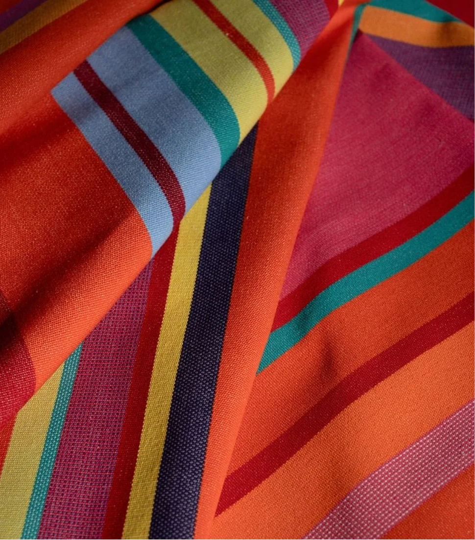 Close up of Les Toiles du Soleil 100% cotton fabric roll of Bonbon Plume - a vibrant orange, pink, and red, accented with anise tones and fine blue striped fabric.