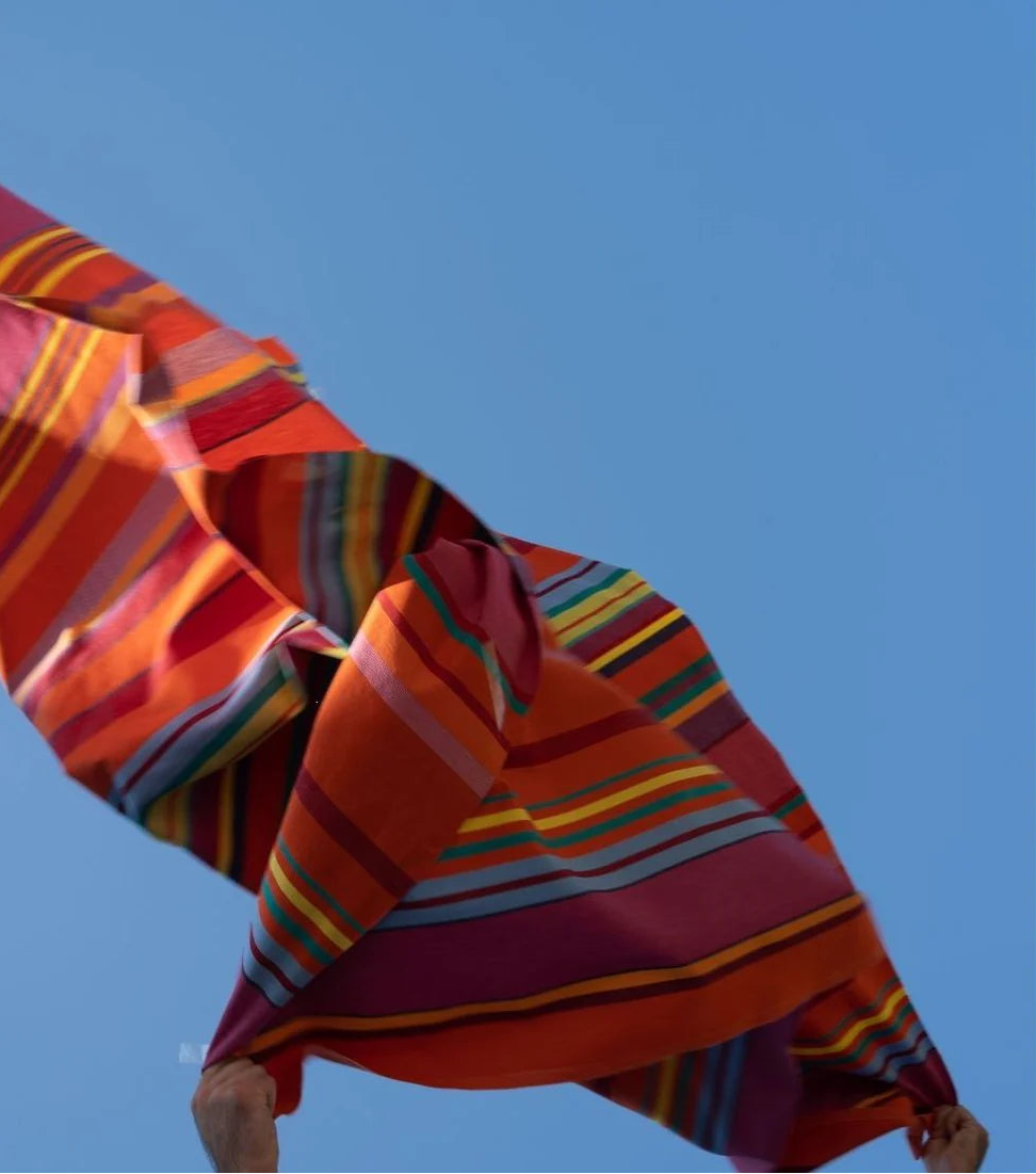 Lifestyle shot of Les Toiles du Soleil 100% cotton fabric Bonbon Plume - a vibrant orange, pink, and red, accented with anise tones and fine blue striped fabric. The fabric is thrown in the air by a person with a blue sky background.