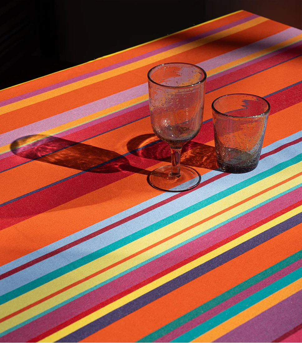 Colourful Les Toiles du Soleil cotton tablecloth in Bonbon Plume pattern, featuring orange, pink, red, anise, and fine blue stripes, styled with 2 drinking glasses