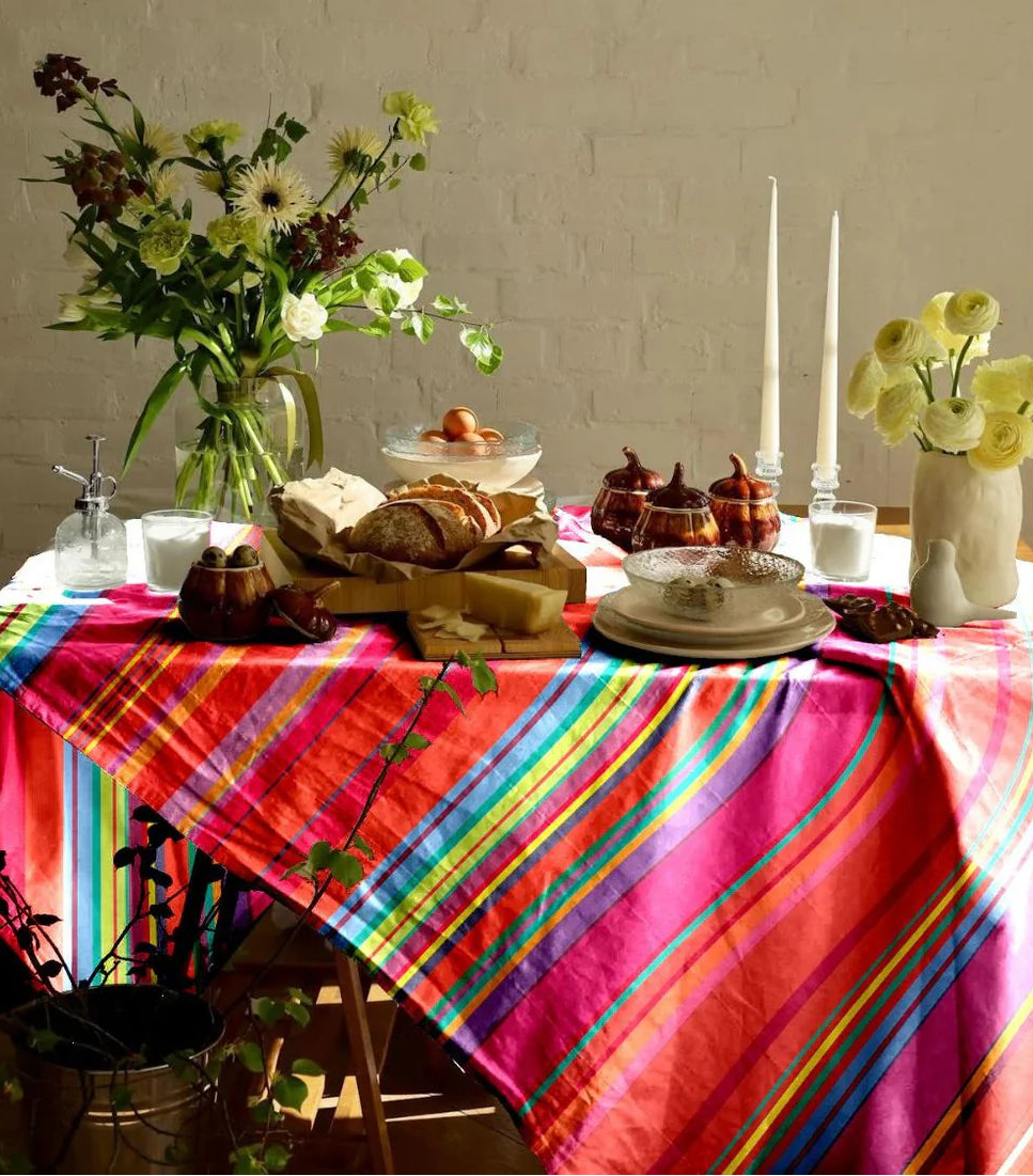 Colorful Les Toiles du Soleil cotton tablecloth in Bonbon Plume pattern, featuring orange, pink, red, anise, and fine blue stripes, styled with food and flowers on top.