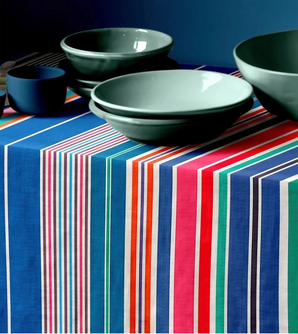 Tablecloth setting of cotton fabric range-Aigue Marine, which is a multi coloured striped fabric made by Les toiles du Soleil. It showcases a variety of shades of blue, pink, red turquoise, greens and other vibrant colours. There are green and blue bowls placed on top to create a lifestyle dining setting shot