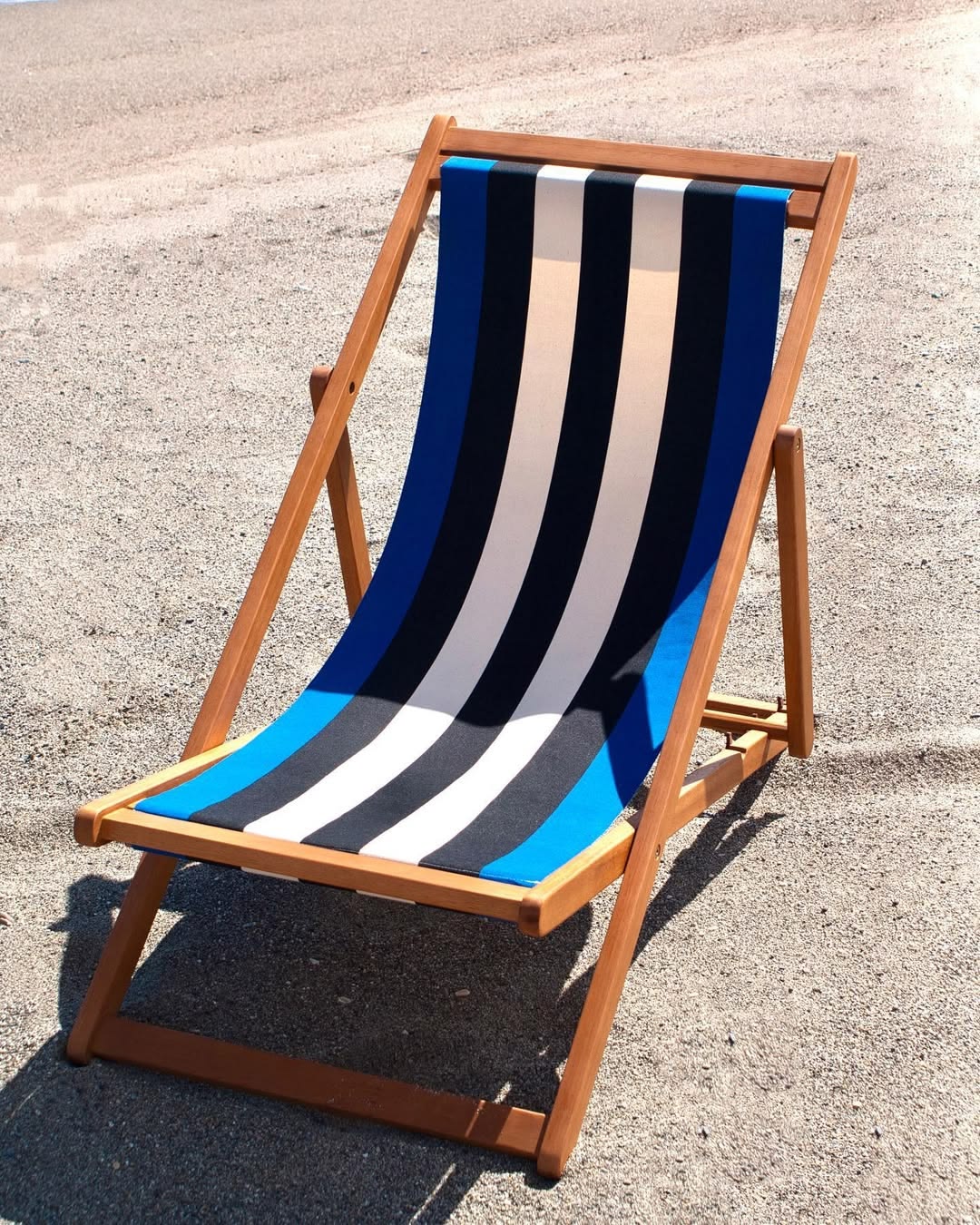 Wooden deckchair on beach showcasing carthage Roy fabric. A bold cotton fabric featuring black and off- white, natural coloured stripes in between marine blue edges. Made by Les Toiles du Soleil