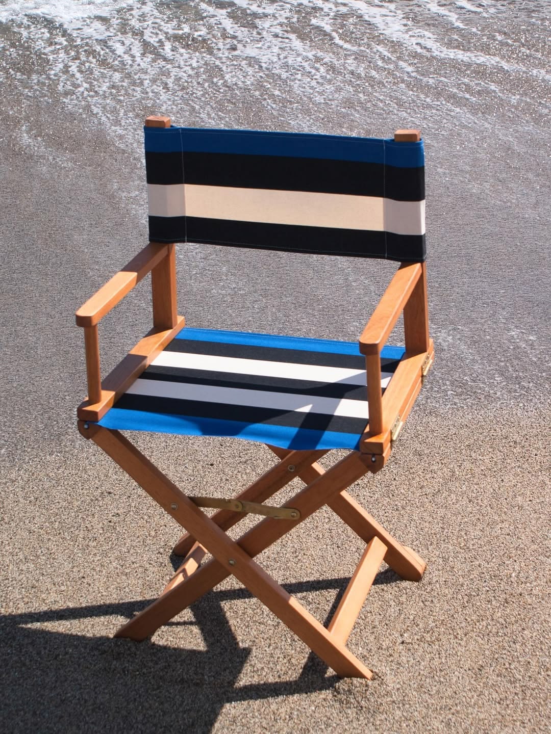Wooden directors chair on beach showcasing carthage Roy fabric. A bold cotton fabric featuring black and off- white, natural coloured stripes in between marine blue edges. Made by Les Toiles du Soleil
