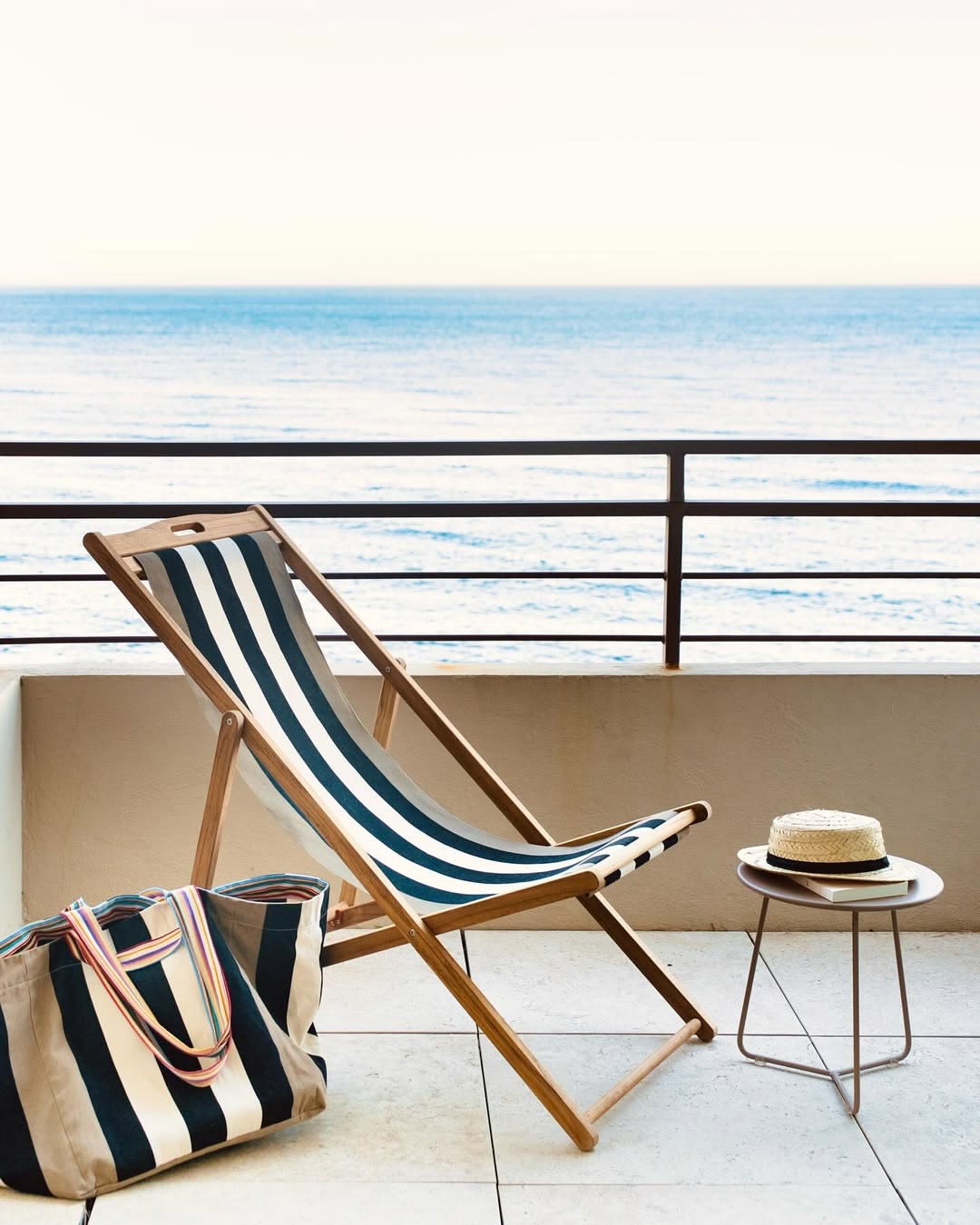 Lifestyle shot on a balcony overlooking the sea. There is a wooden deckchair and large carry bag with small table with a straw hat and book. The fabric for the deckchair and bag is cotton fabric featuring bold black, ecru stripes in-between taupe coloured stripes on the edges by Les Toiles du Soleil