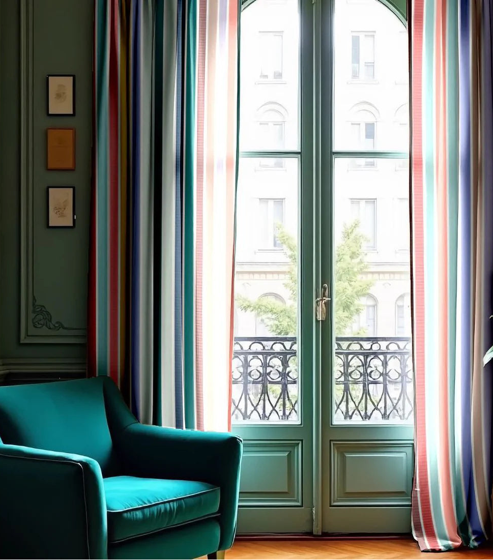 Teal armchair in a room with large windows and colorful curtains made from the Eze range of Les Toiles du soleil cotton fabric. Multi coloured striped  pastels, blues, purples, mint green, and other joyous hues