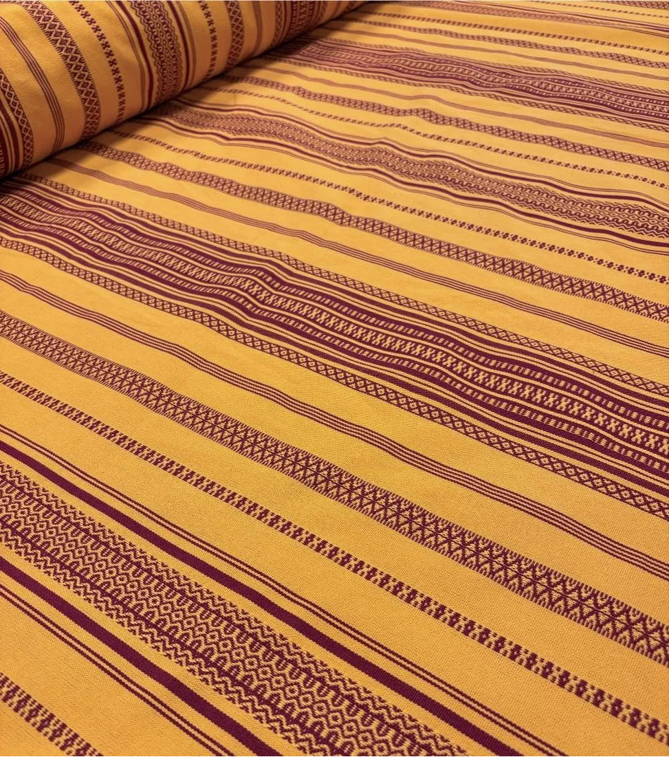 Close up of 100% cotton fabric roll of Grenade Safran by Les Toiles du Soleil. Intricate woven stripes of burgundy and saffron textured fabric