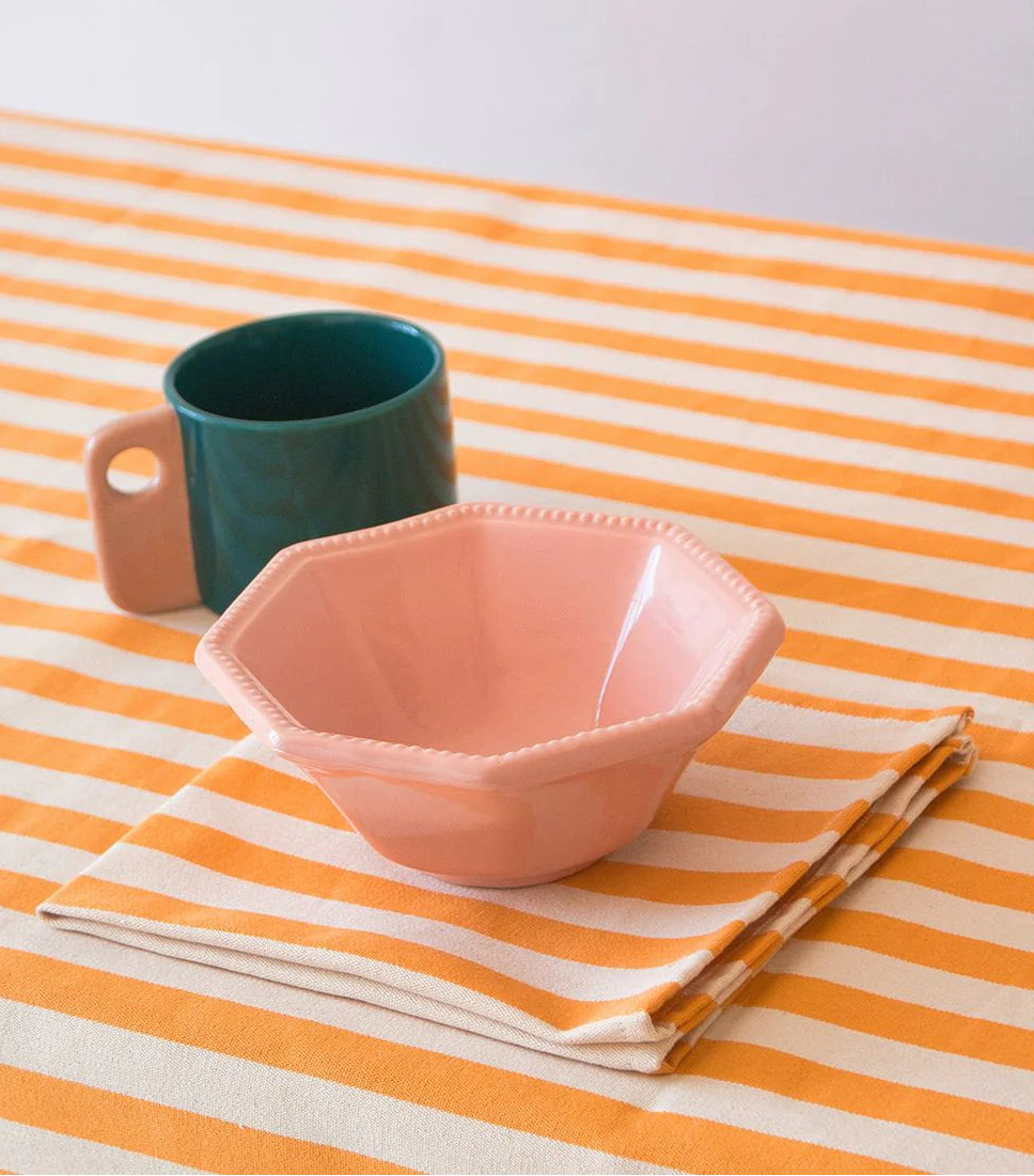 Pink bowl, green mug, and folded napkins and ecru and saffron coloured striped cloth on a matching striped tablecloth. Mafe by Les Toiles du Soleil - Isola Bella