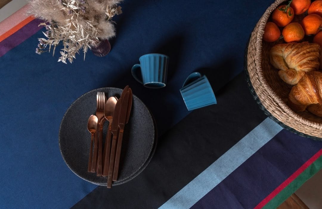 Close up of fabric Tablecloth of Lucas a fabric bold striped Les Toiles du Soleil woven fabric of deep purple, blush pink, burnt orange, petrol blue, black and forest green. The tablecloth has plates, cutlery and a basket of fruit and pastries on it