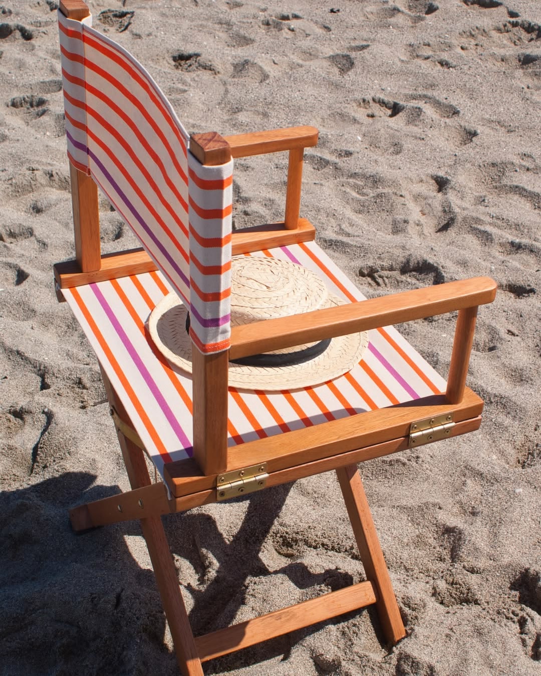 Straw hat on a directors chair on a beach made from vibrant thin sailor stripes of orange and fuchsia on a 43cm 100% cotton fabric called Marin Orange by Les Toiles du Soleil.