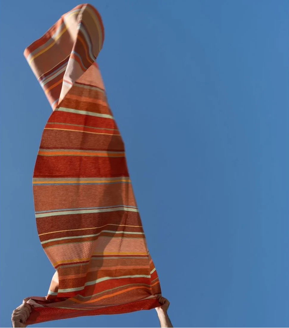 Striped fabric in the air with a blue sky background. The fabric is multi colours - ight stone and terracotta, soft greens, subtle rusts and the patinated reds.