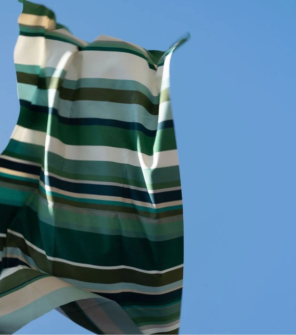 Place aux Herbes - a 100% cotton fabric roll which is a vibrant striped tapestry: fresh greens, almond, olive and taupe, fabric being thrown in the air with a sky blue background