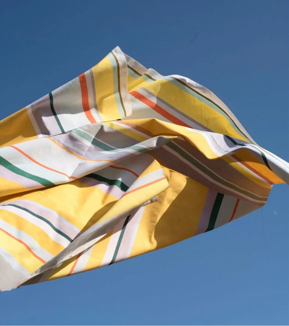  Lifestyle shot of Plein Soleil multi coloured yellow, grey, and sun toned striped Les Toiles du Soleil 100% cotton fabric being thrown in the air with a sky blue background
