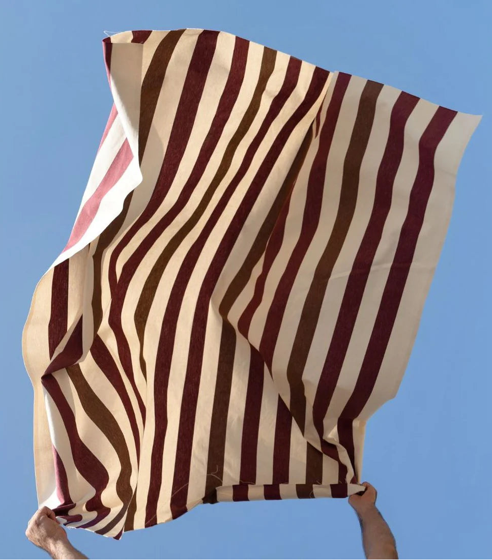 Les Toiles du Soleil Raspail Prunelle 100% cotton striped fabric of plum, browns and white thrown in the air with a blue background
