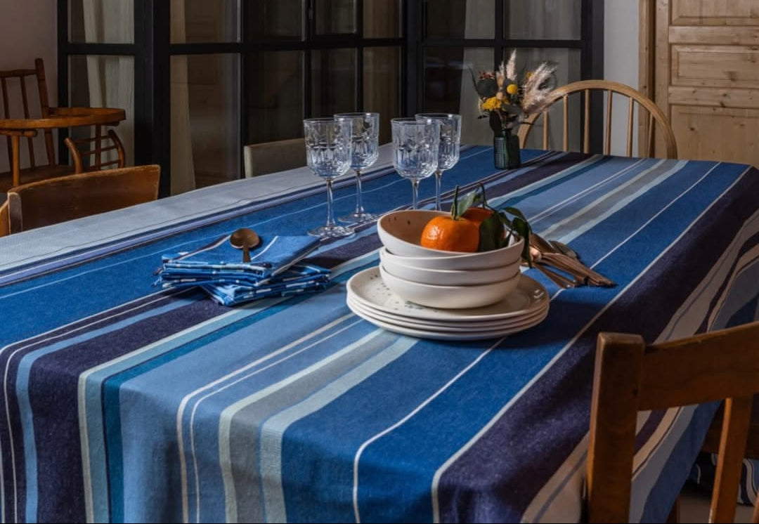 Dining table setting with Rue du Bac Bleu cotton fabric of multiple shades of striped blue. Plates and glasses set on table cloth with glass windows in background. 