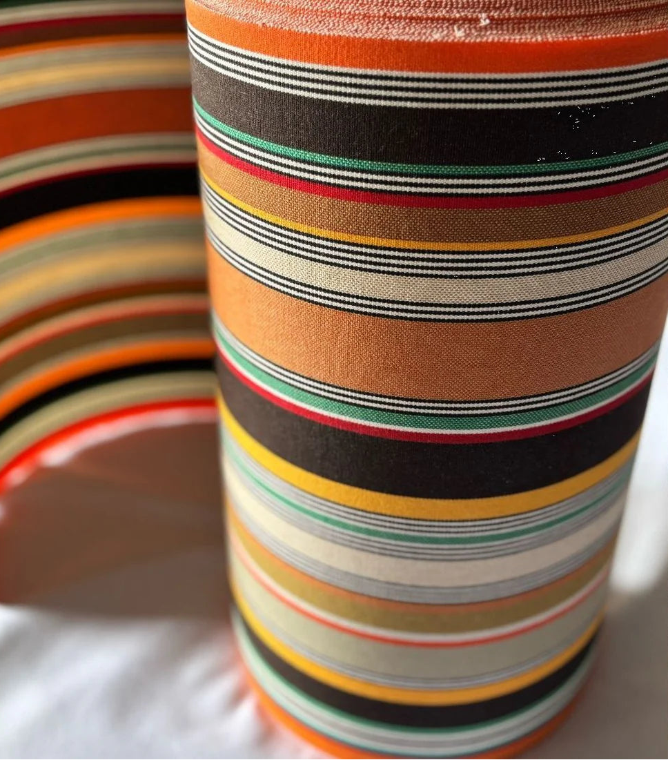 Close up of cotton fabric roll featuring densely woven stripes of orange, dark brown, golden yellow, teal, sand, olive, red and cream, with fine black and white pin lines throughout and bright orange selvedge edges.