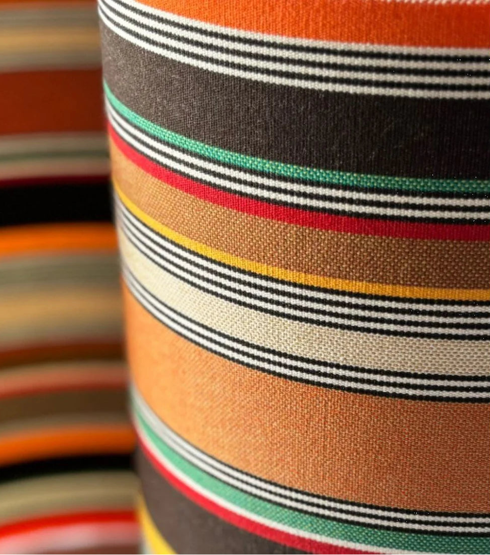 Close up of cotton fabric roll featuring densely woven stripes of orange, dark brown, golden yellow, teal, sand, olive, red and cream, with fine black and white pin lines throughout and bright orange selvedge edges.