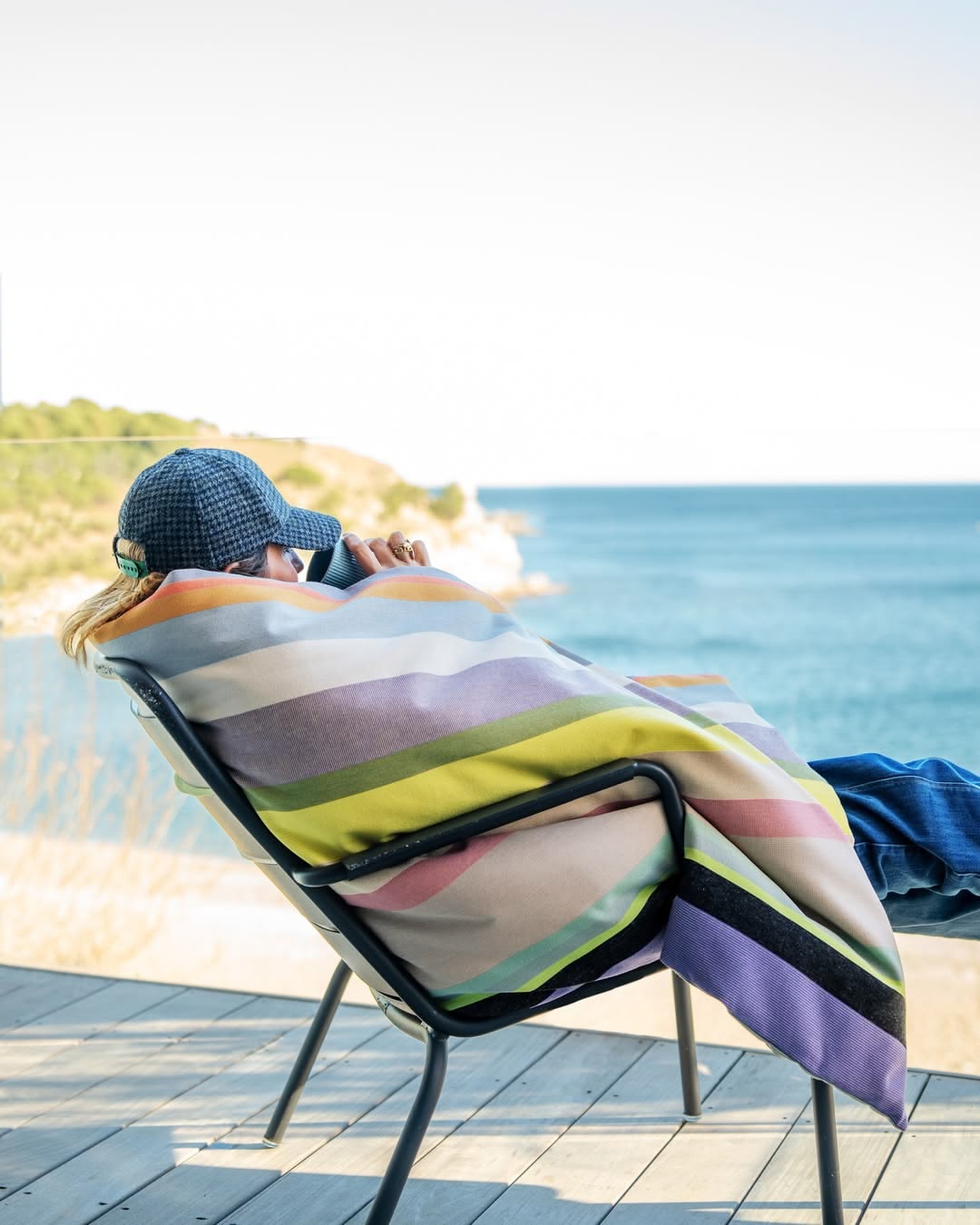 Person sitting on a chair with a colourful multi striped throw draped over it by the ocean.