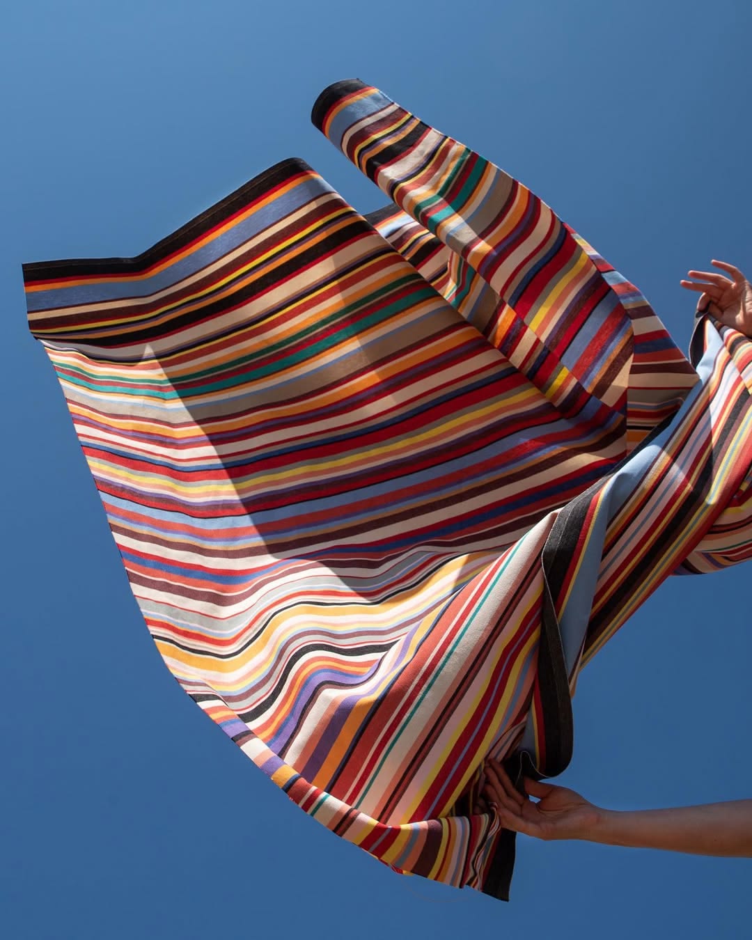 Les Toiles du Soleil's Tom Multi Outdoor Sunbrella Fabric, featuring multi coloured stripes in the air with a blue sky background