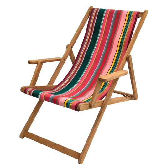 On a white background a wooden teak folding deck chair with arms featuring a vibrant striped, pink, red, green fabric sling made by Les Toiles du Soleil.