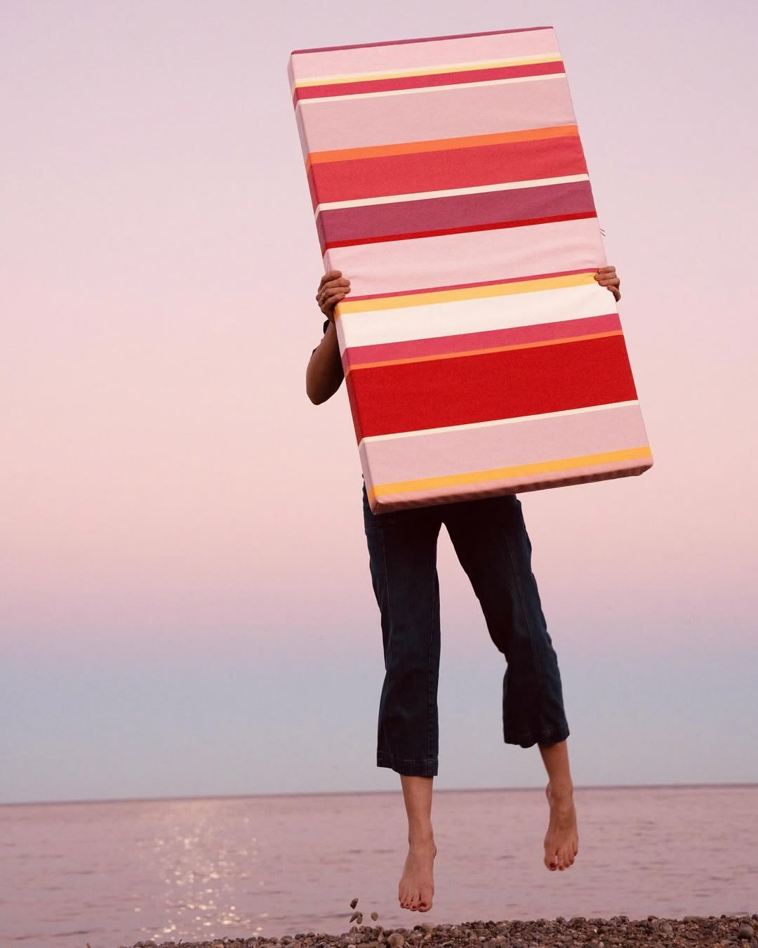 A person on the beach holding a large slim cushion made from Zinnia Les Toiles du Soleil Outdoor Sunbrella Fabric. Multi striped with rose, pinks, reds and yellows