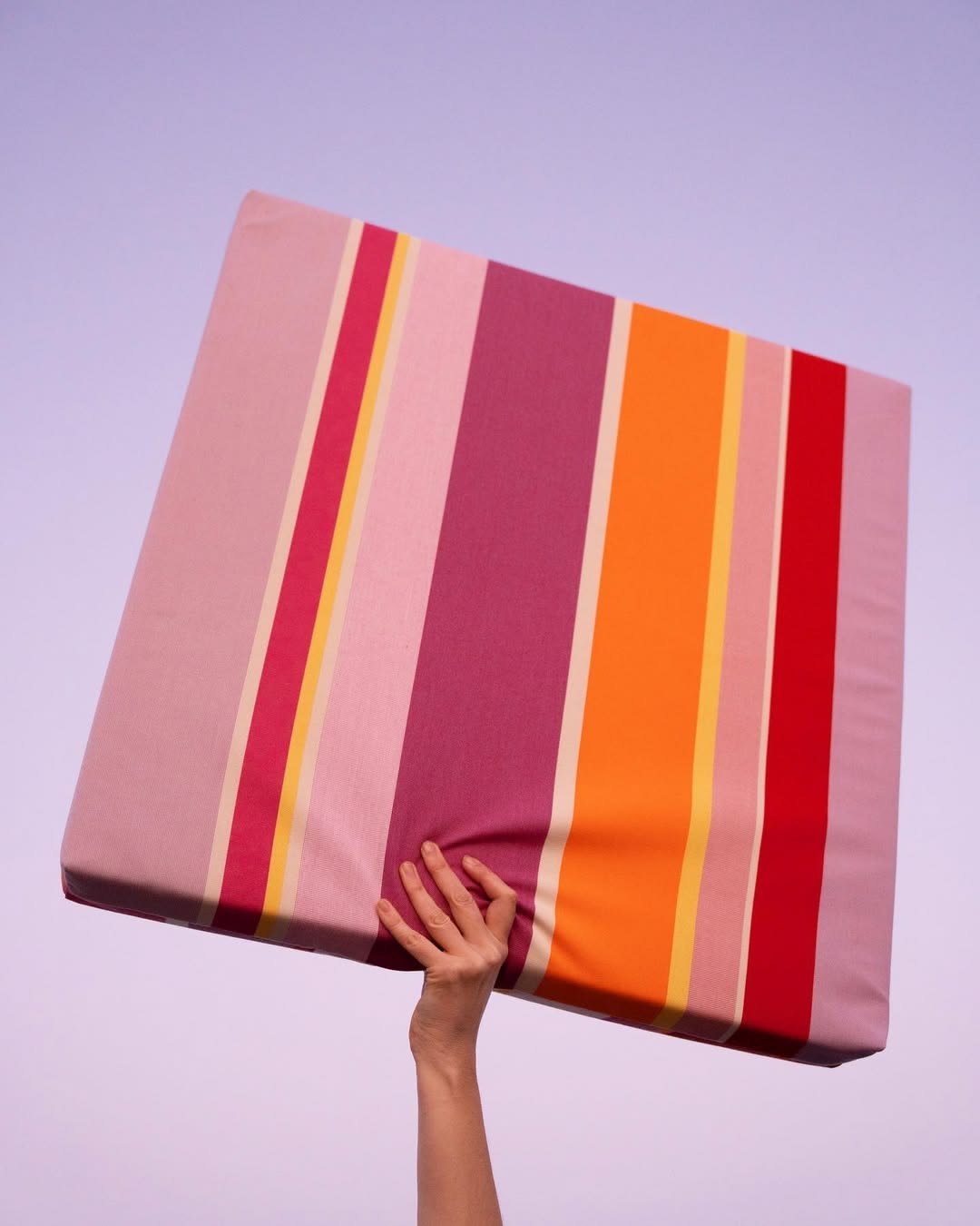A person holding a slim cushion in the air made from Zinnia Les Toiles du Soleil Outdoor Sunbrella Fabric. Multi striped with rose, pinks, reds and yellows. 